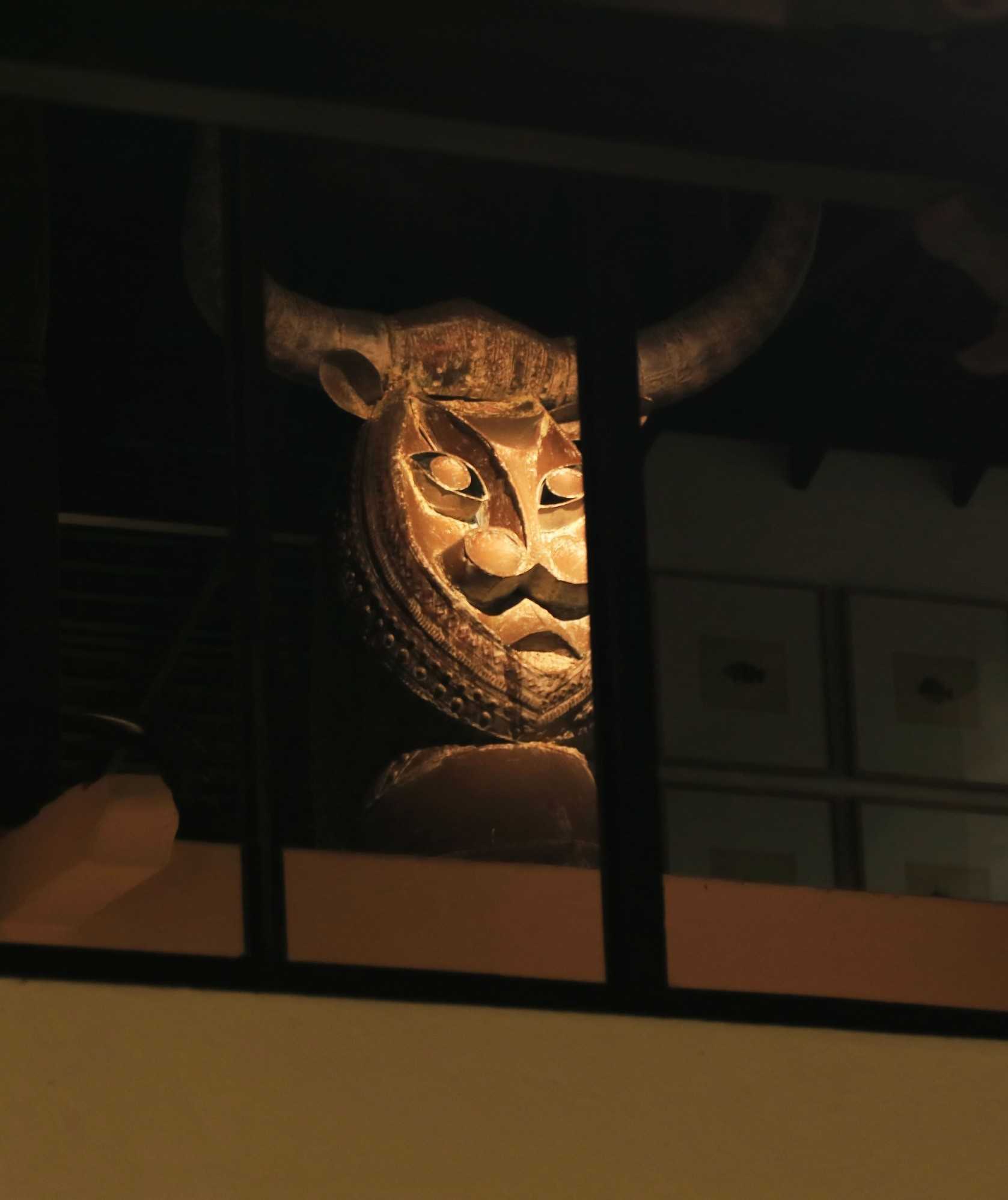 A carved wooden bull's head with long curved horns, lit from the side, seen through dark window bars.