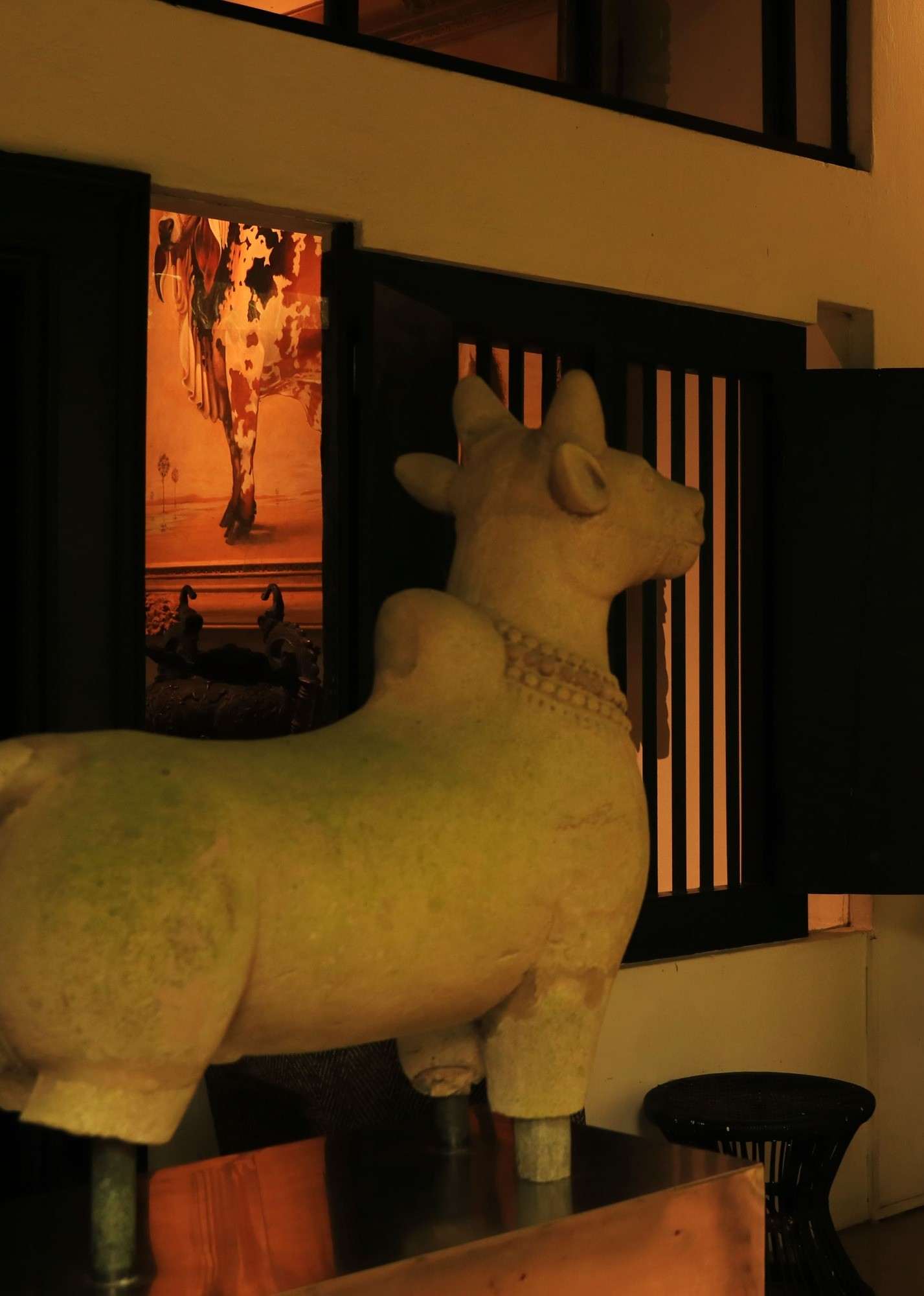 A pale sculpted bull with a garland about its neck, standing in front of a window with black bars.