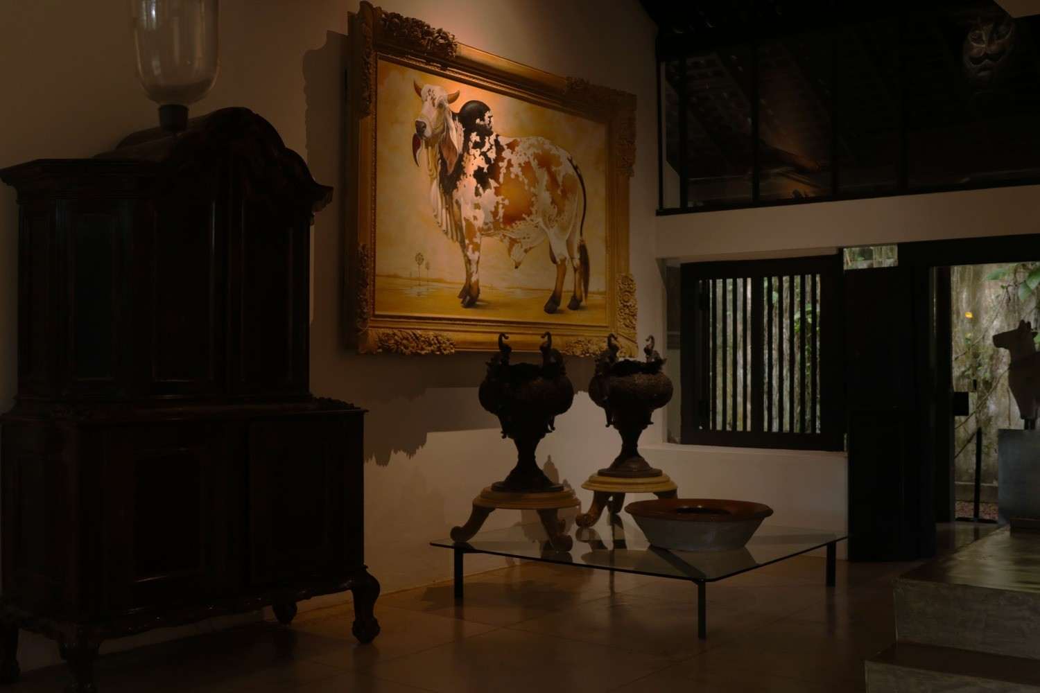 A hall with an old dark cabinet, a gilt-framed painting of a cow, a pair of urns on a glass table and a latticed window.