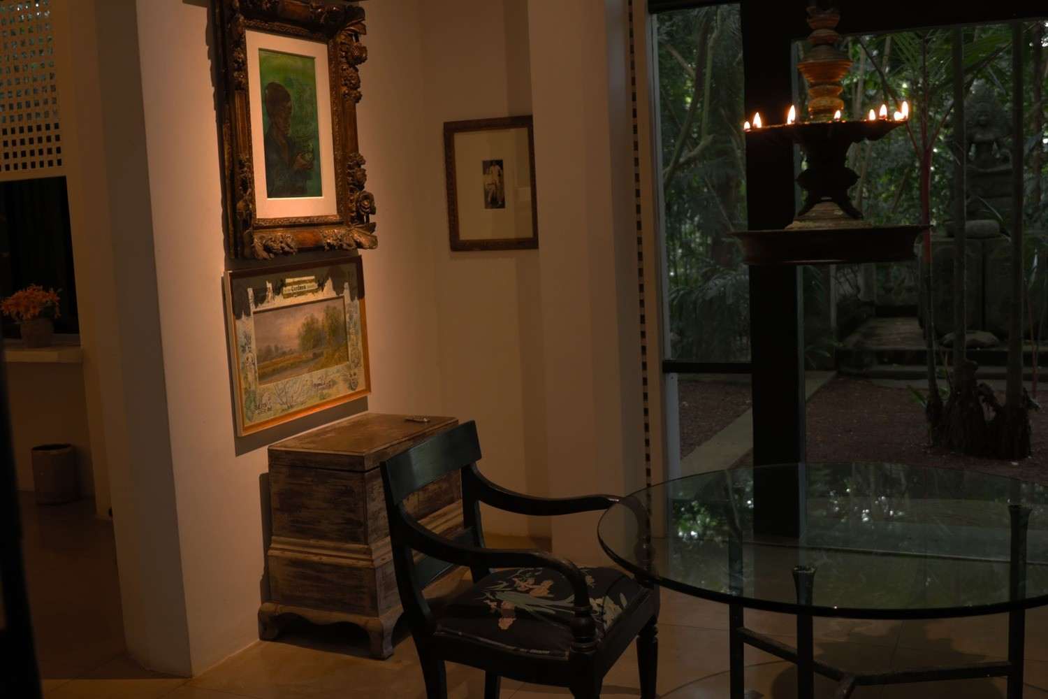A white room with framed paintings and a lamp, an old chest, and a wide latticed opening onto the garden.