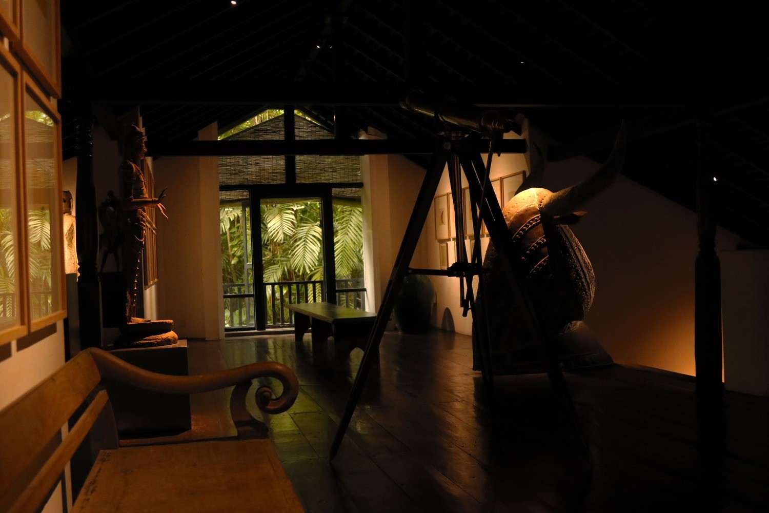 A dim hall of dark timber and white walls, a carved figure lit by a lamp, the garden green through the far opening.