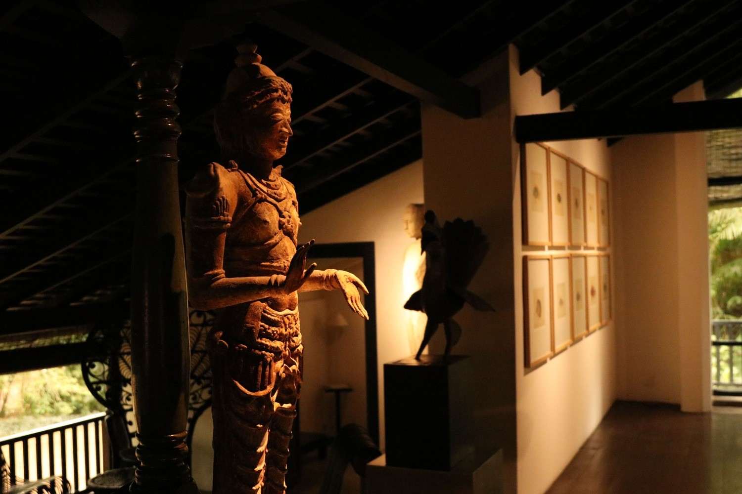 A dim, high-ceilinged hall of dark timber: a carved figure by a column, framed drawings on a white wall, and the garden bright through an opening.