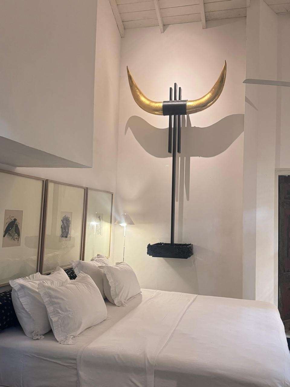 A sculpture of a pair of golden bull horns on a dark post, mounted on the white wall above the bed.