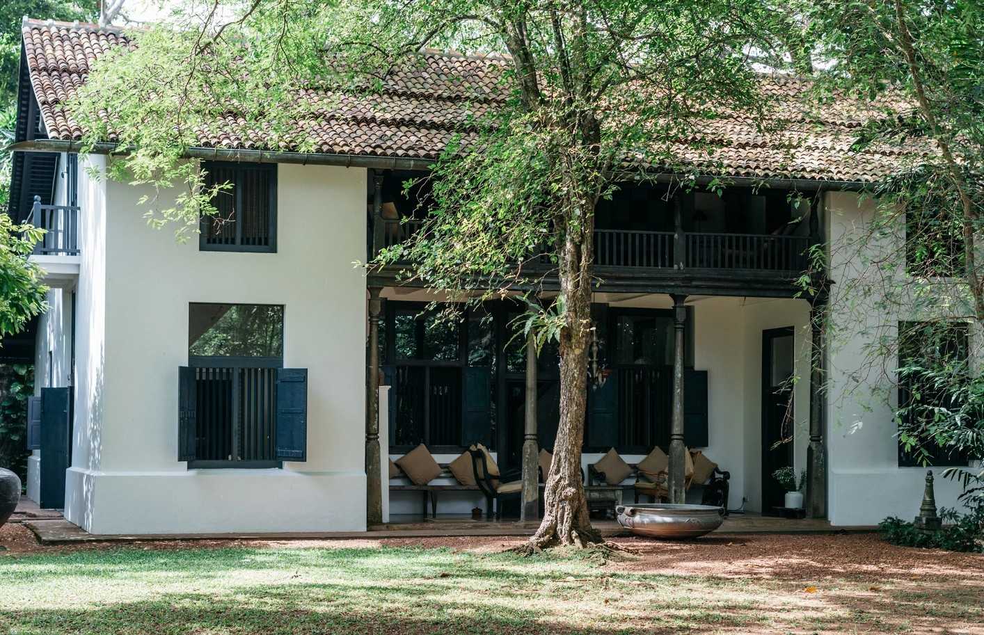 The whitewashed two-storey house with dark green shutters and a tiled roof, behind a large tree on the lawn.