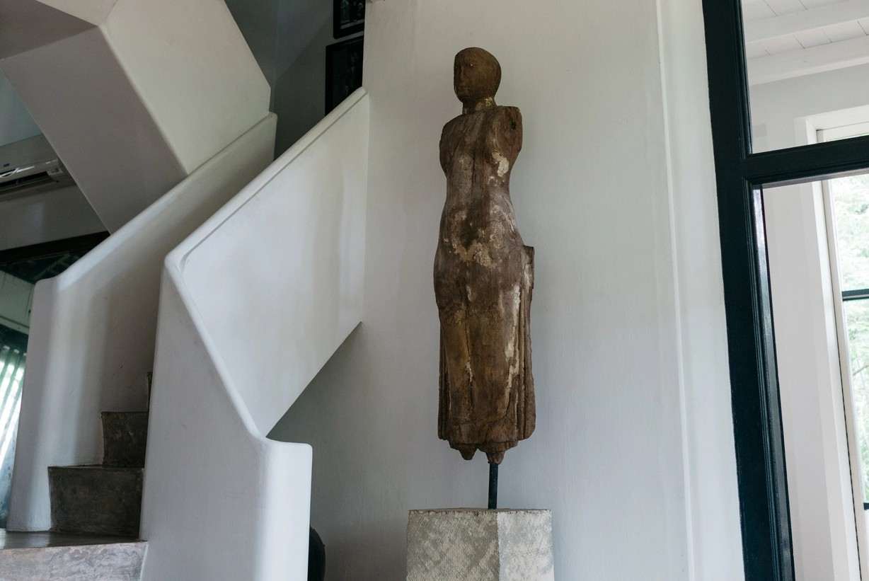 A white staircase rising in plain, sculptural steps, a tall carved wooden figure on a stone plinth at its foot, a glass lantern hanging above.