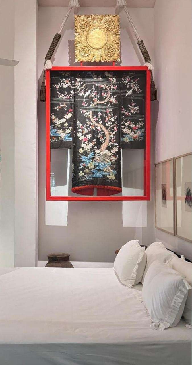 An embroidered black kimono with birds and branches, framed in red on a white wall, above the bed.