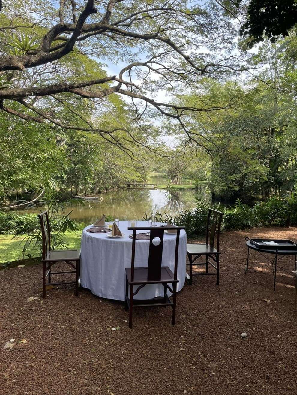 A table for two with a white cloth on the laterite ground by the lake, under the trees.