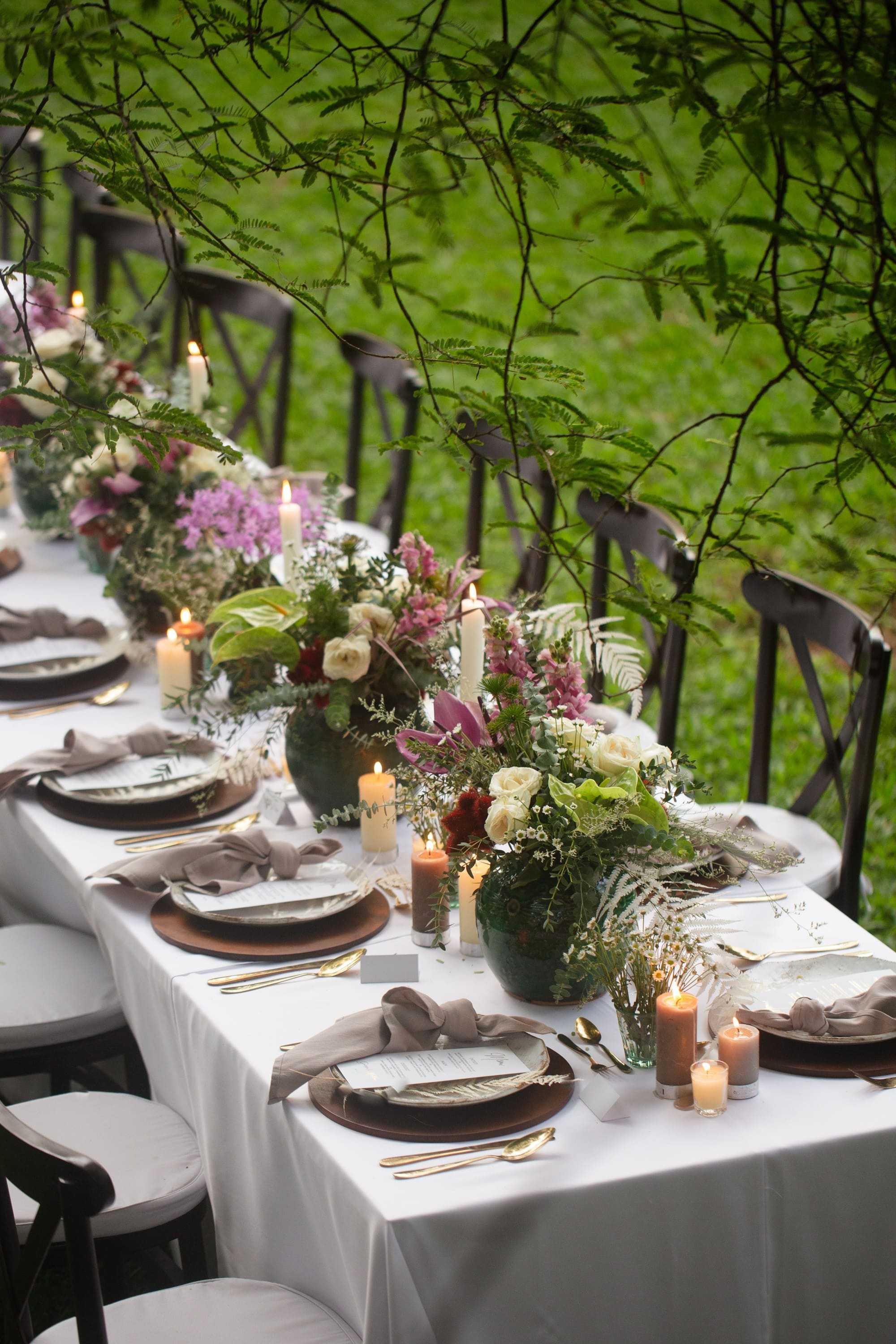 A long table laid in the garden: white cloth, gold-rimmed plates, candles and low arrangements of pink and white flowers.